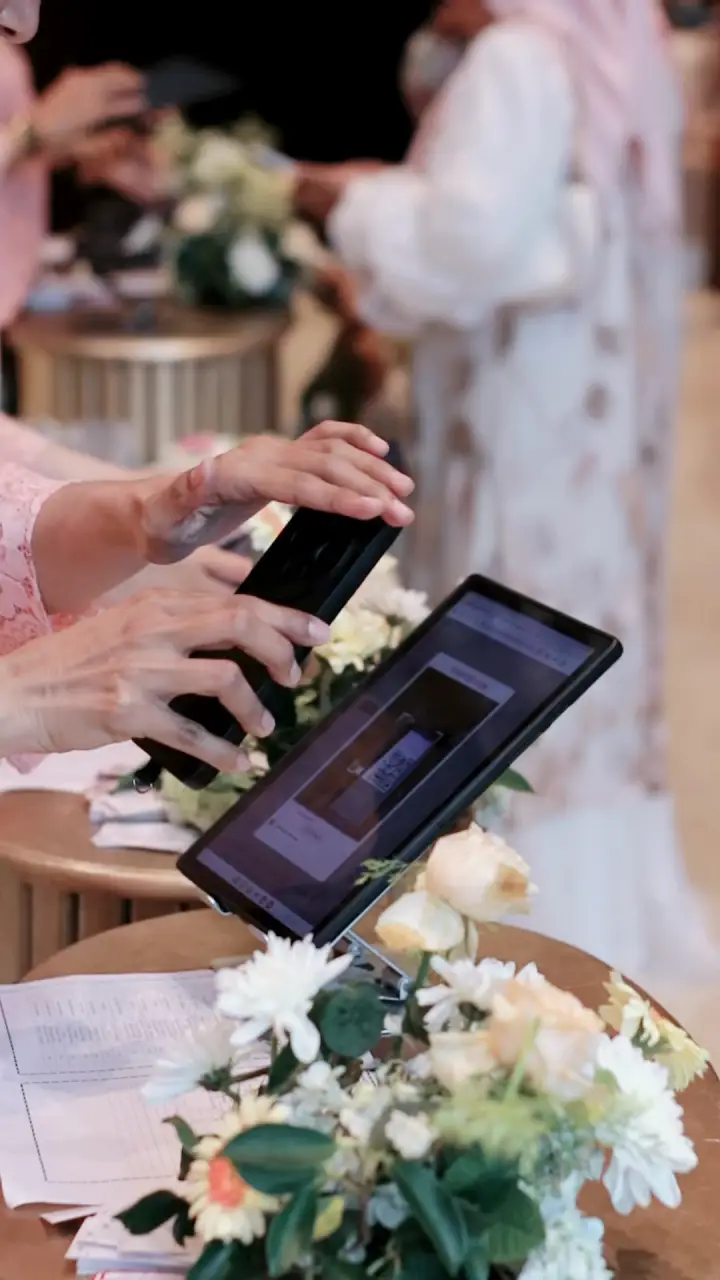 Tamu menunjukkan QR di ponsel ke tablet di meja penerimaan, lalu penerima tamu berkebaya mencatat kehadiran.